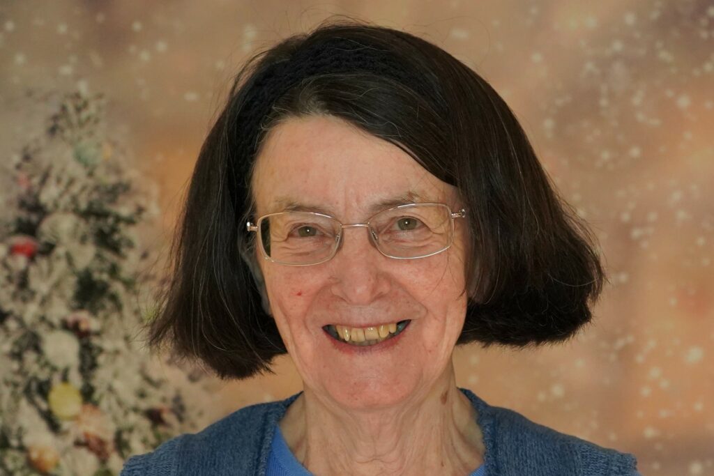 Headshot of Marg, a young-looking woman in her 70s with white skin and glasses, dark hair in a straight bob just above her neck line. There is the edge of a Christmas tree to the left of her and fake snow on the peach-coloured background.