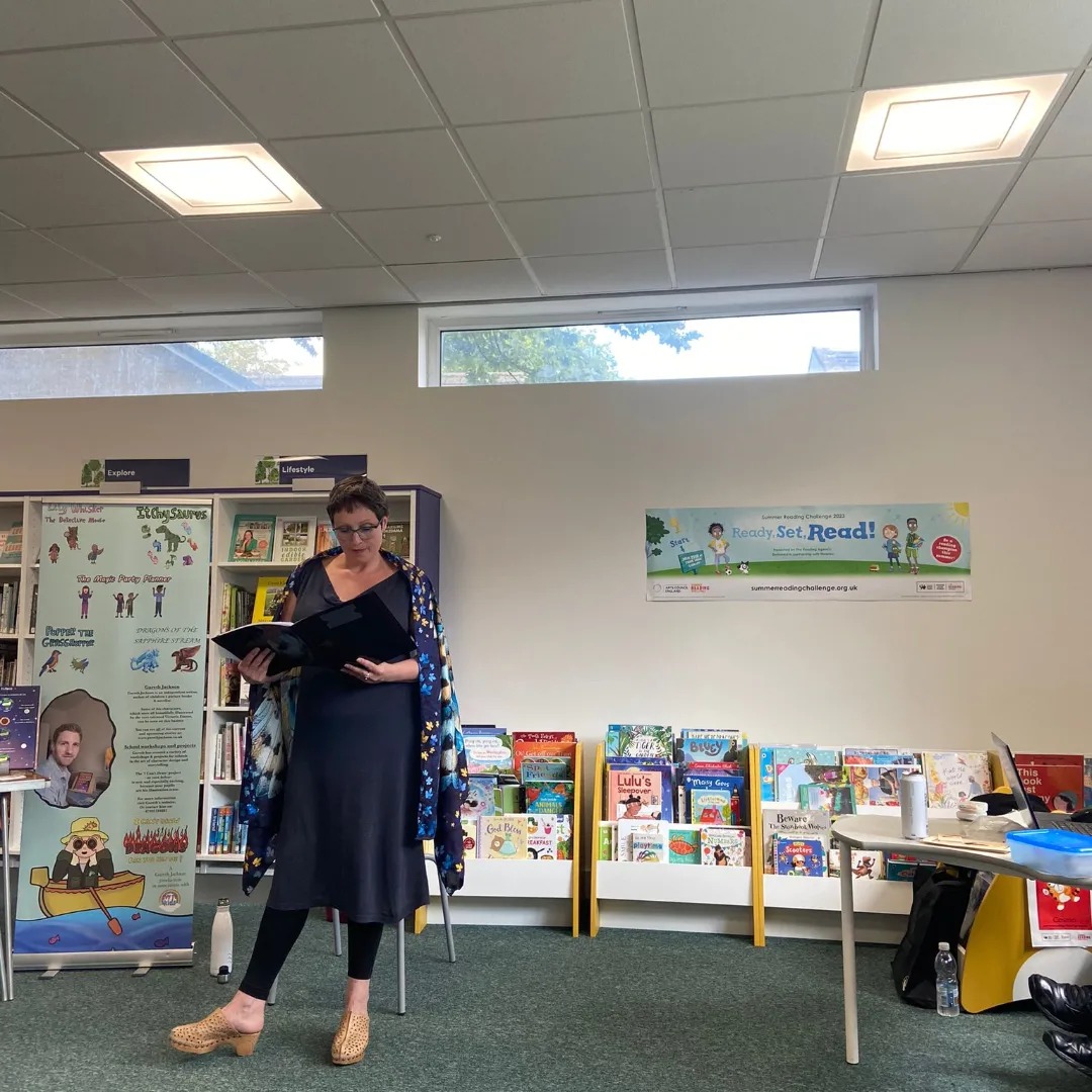 Library with children's books in stands, a table and banner aboit Gareth Jackson's writing. Becky with short dark hair flecked with grey and glasses, wearing long blue dress, blue patterned shawl and beige clogs.
