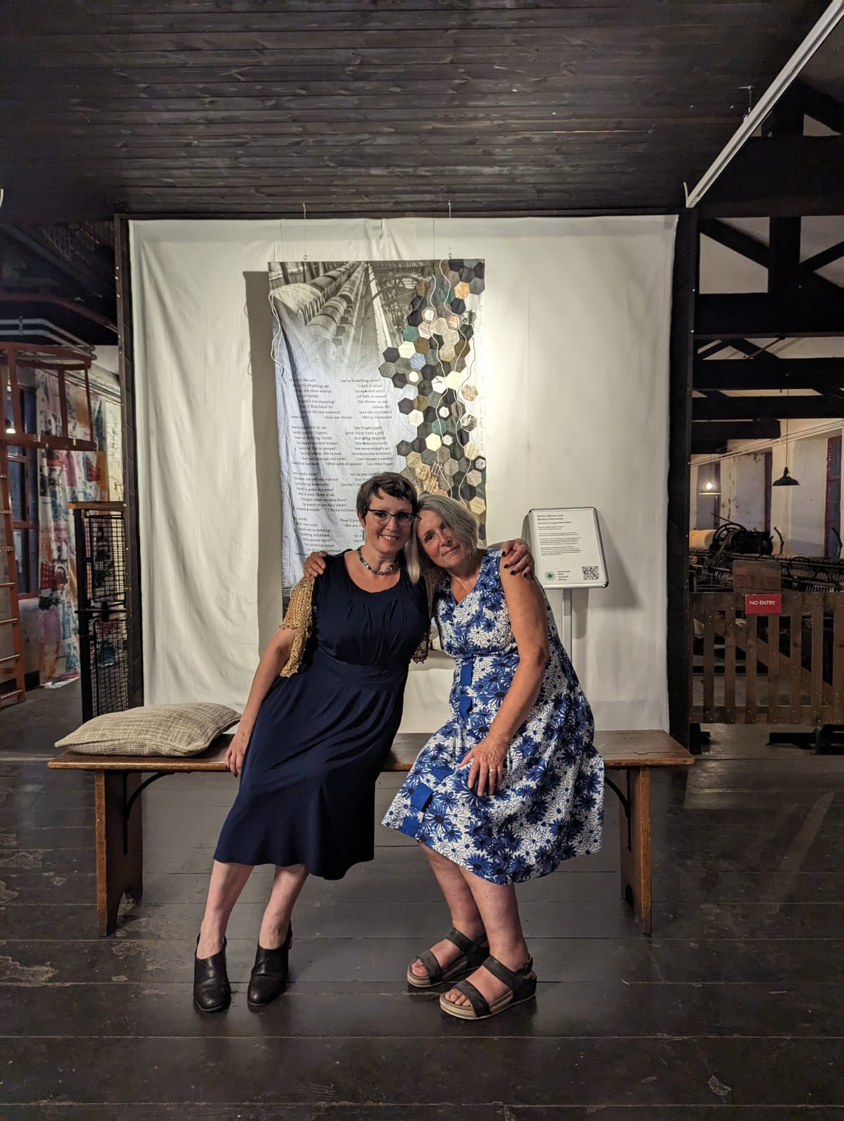Any work that wanted doing exhibition. Becky Cherriman and Becky Moore sit on a bench in front of the work facing the camera with their arms around one another’s shoulders. Becky C is on the left wearing glasses and a blue and silver necklace in long purple dress and gold shrug. Becky M is wearing a blue and white floral dress featuring a weave of blue cloth up the front centre line to the waist. Behind them, you can see ¾ of the textile hanging, from the top. There is a photograph of a loom at the top. On the right a patchwork of hexagons in different materials and muted colours interspersed by patches of ephemera. The poem is laid out on the left and centre of the piece in a regular pattern with gaps and back slashes between phrases. The writing is unclear.