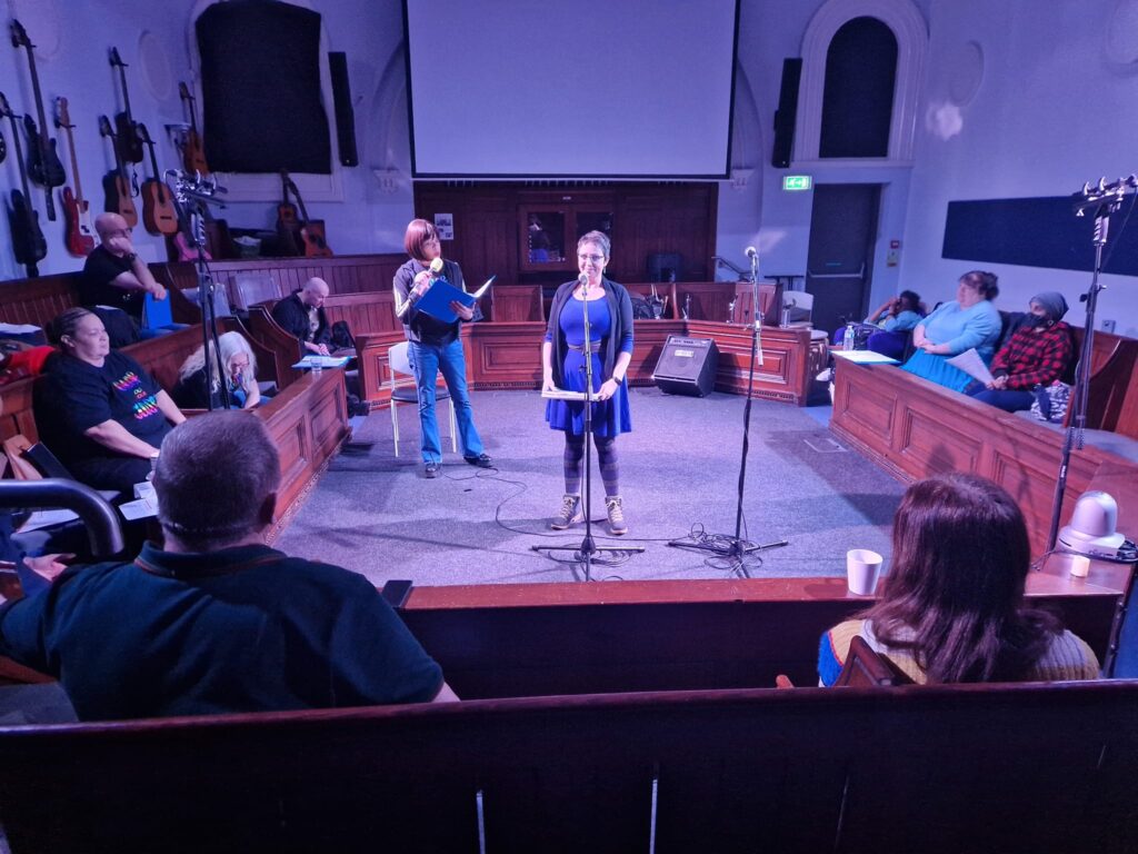 Becky in purple dress performing at Chapel FM, a church with host Michelle Scally Clarke behind and audience in the pews.